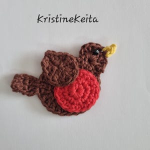 Könnte beinhalten: Ein handgehäkeltes Vogel-Design in Braun-, Rot- und Gelbtönen. Der Vogel hat eine rote Brust, braune Flügel und einen Körper sowie einen gelben Schnabel. Der Text "Kristinekeita" ist über dem Vogel geschrieben.