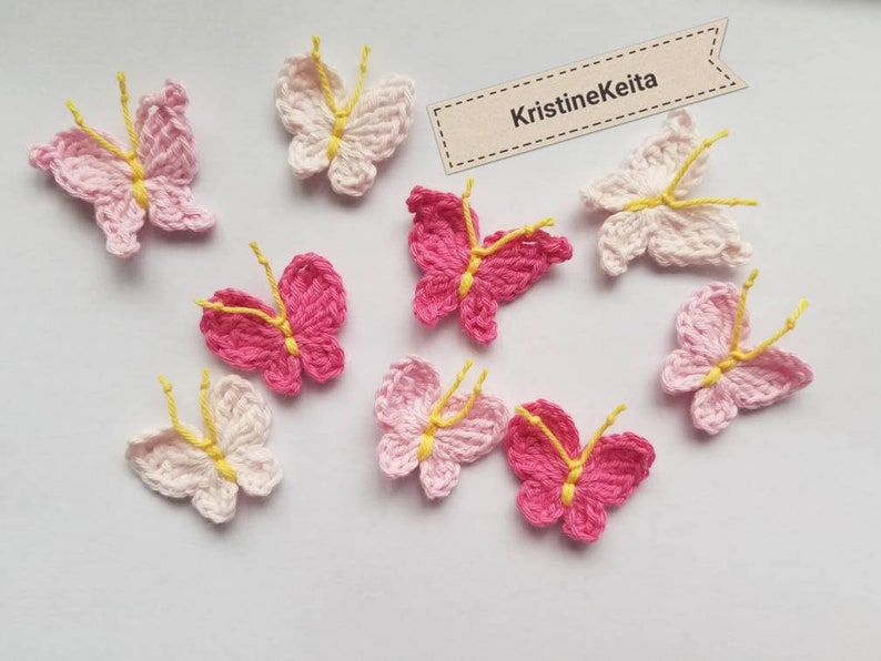 May include: A collection of handmade crochet butterflies in various shades of pink and white, with yellow accents. The butterflies are arranged on a light background. The image includes a tag with the text "KristineKeita".