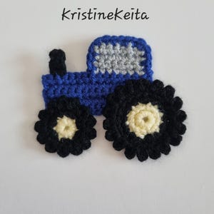 May include: A blue and grey crocheted tractor appliqué with black and cream wheels and a black exhaust pipe. The text KristineKeita is printed above the tractor. This textile craft piece features detailed yarn stitching throughout the design.