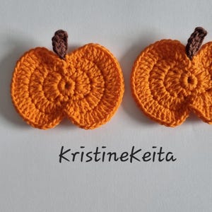 May include: Two orange crochet pumpkin appliques with brown stems, featuring a textured circular stitch pattern. The text KristineKeita is printed in black below the two pumpkins, which are arranged side by side on a plain white surface.