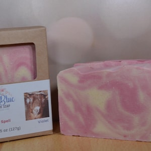 May include: A bar of Irish Blue Goat Milk Soap, Love Spell scent, with a violet hue and a swirled yellow pattern. The soap is next to its packaging, which includes the brand name, a goat image, and the net weight of 4.5 oz (127g).