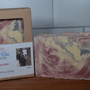 May include: A rectangular bar of goat milk soap with a marbled pattern of red, cream, and dark gray, packaged in a brown cardboard box with a window. The label features the text Irish Blue Goat Milk Soap, Amazing Style, and a photo of a goat.