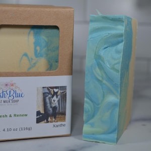 May include: A bar of Irish Blue goat milk soap with blue and yellow swirls, next to its packaging. The packaging is a brown box with a window showing the soap. The label reads "Refresh & Renew" and "Net Wt. 4.10 oz (116g)".
