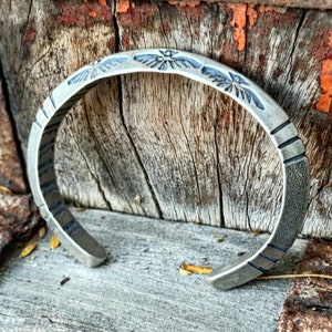 May include: A silver bracelet with a textured finish and a design of stylized birds. The bracelet is open and has a slightly curved shape.