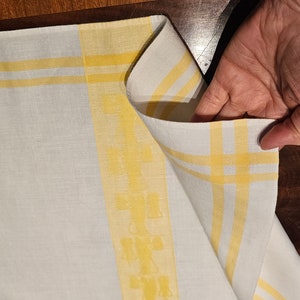 May include: White linen tablecloth with yellow stripes and a grid pattern. The fabric is folded, revealing the underside. The tablecloth is likely for a dining table or kitchen use.