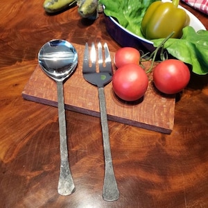 May include: A stainless steel serving spoon and fork with hammered handles rest on a small wooden cutting board. Red tomatoes and a green bell pepper are nearby, suggesting a culinary theme.