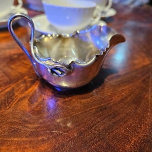May include: A silver creamer with a handle and a ruffled edge. The creamer has a decorative floral accent on the side. The creamer is sitting on a dark wooden surface. Teacups are visible in the background.