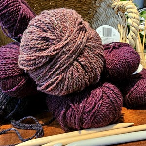 Op de afbeelding: Verschillende bollen paars en bruin garen, met breinaalden en een mand op de achtergrond. Het garen ligt op een houten oppervlak, wat wijst op een knutselthema.