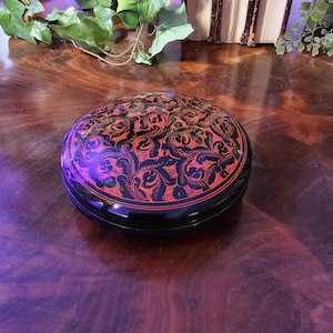 Könnte beinhalten: Eine runde, schwarz lackierte Box mit einem roten Blumendesign auf dem Deckel. Die Box steht auf einer dunklen Holzoberfläche. Es ist ein dekorativer Aufbewahrungsbehälter.