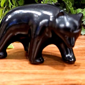 May include: A glossy black bear figurine. The bear is depicted in a walking pose, with its head slightly raised. The figurine is made of a smooth, reflective material, and sits on a wooden surface.
