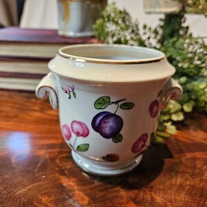 Op de afbeelding: Een witte keramische plantenbak met gouden rand en handvatten. De plantenbak is versierd met paarse pruimen en roze bloemen met groene bladeren. De plantenbak staat op een houten oppervlak.