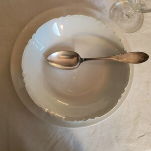 May include: A white porcelain bowl with a scalloped edge sits atop a larger white plate. A silver spoon with ornate detailing rests in the bowl. The scene is set on a white tablecloth, with a glass visible in the background.