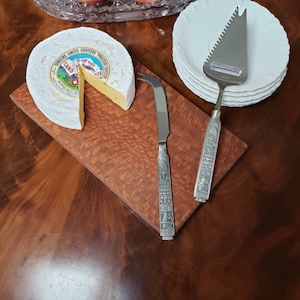 May include: A wheel of Swiss Gruyere cheese with a wedge cut out, displayed on a wooden cutting board. A cheese knife and a cheese slicer with decorative handles are next to the cheese. White plates are stacked nearby.