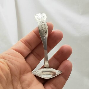 May include: A small, ornate silver spoon with a decorative handle and a flared, shallow bowl. The handle features engraved floral patterns. The spoon is held in a hand against a white background, showcasing its intricate design and craftsmanship.