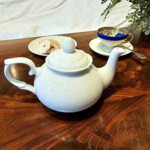 May include: A white ceramic teapot with a rounded body, a curved handle, and a lid with a small knob. A white teacup with a blue and gold rim sits on a saucer, with a spoon. A plate of cookies is also present.