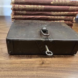 May include: A weathered, rectangular metal box with a hinged lid, a keyhole, and a key. The box has a handle and is riveted at the corners. A stack of antique books with red covers is in the background.