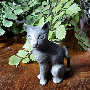 May include: A gray cat figurine in a seated position with its tail curled. The cat has detailed features and is placed on a wooden surface with green foliage in the background. The figurine is a decorative item.