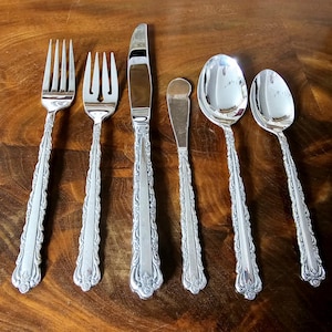 以下が含まれることがあります： 2 本のフォーク、ナイフ、バターナイフ、2 本のスプーンを含む、銀色のカトラリー 6 点セット。ハンドルには詳細な花の模様があります。カトラリーは、濃い木製の表面に配置されています。