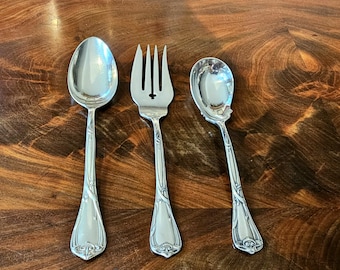 May include: Three pieces of antique silverware: a spoon, a fork, and a smaller spoon. The silverware is silver-coloured with ornate floral designs on the handles. The fork has a decorative cross-like design at the top.