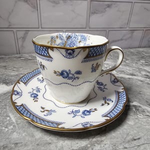 May include: A white teacup and saucer set with blue floral and geometric patterns, accented with gold trim. The teacup has a curved handle and a scalloped rim. The saucer mirrors the cup's design, creating a classic tea set.