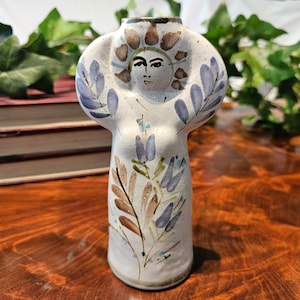 Puede incluir: Un jarrón de cerámica con un diseño de rostro y flores. El jarrón es de color blanco roto con detalles en azul, marrón y verde. La cara tiene ojos oscuros y una expresión neutra. El jarrón mide unos 25 cm de altura.