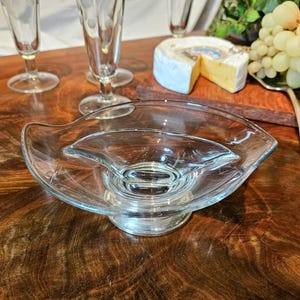 Op de afbeelding: Een heldere glazen serveerschaal met een uniek, gebogen ontwerp, rustend op een zilverkleurige basis. De schaal heeft een centraal, verhoogd gedeelte en een brede, ondiepe buitenrand. Andere glaswerk en voedsel zijn op de achtergrond.