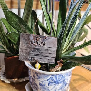 May include: A metal garden sign with the words "MY GARDEN" embossed, suspended in front of a potted plant. The sign is attached to a copper wire. A small, yellow and white bead hangs from the bottom of the sign. The plant has long, green leaves.
