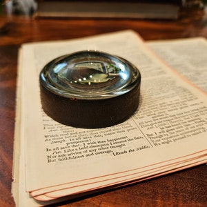 May include: A clear glass magnifying lens with a dark brown leather base rests on an open book. The book's pages are aged, with visible text. The lens reflects the surrounding environment, creating a vintage aesthetic.