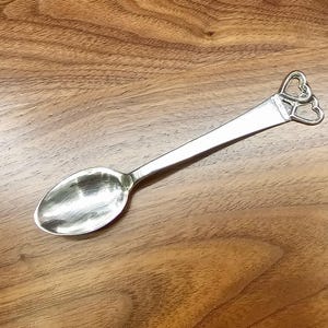 May include: A silver spoon with a heart-shaped design at the handle's end. The spoon has a rounded bowl and a long, slender handle. The handle features two connected heart shapes. The spoon is set against a wooden surface.