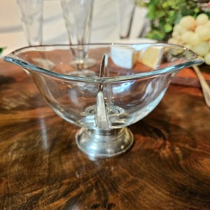 Op de afbeelding: Een heldere glazen serveerschaal met een zilverkleurige voet, verdeeld in twee secties. De schaal staat op een donker houten oppervlak, met op de achtergrond verschillende geëtste champagneflutes. Kaas en druiven zijn ook zichtbaar.