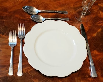 May include: A formal place setting with a white scalloped-edge plate, two forks, two spoons, and a knife. The silverware has decorative detailing. A wine glass is also present. The items are arranged on a dark wood surface.