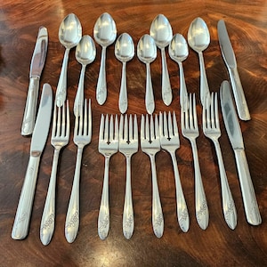 May include: A collection of vintage silverware, including spoons, forks, and knives, arranged on a dark wooden surface. The silverware has a floral design on the handles and a shiny, reflective surface. The set includes various sizes of spoons and forks.