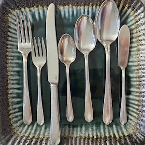 May include: Silverware set including forks, knives, and spoons arranged on a decorative plate. The set includes two forks, a knife, a teaspoon, a tablespoon, a soup spoon, and a butter knife. The silverware has a reflective surface.