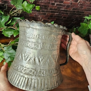 Könnte beinhalten: Ein antiker silberfarbener Metallkrug mit Henkel und gewelltem Rand. Der Krug ist mit aufwendigen geprägten Mustern verziert, darunter geometrische Formen und florale Designs. Der Krug wird von zwei Händen gehalten.