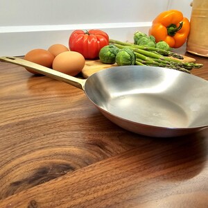 May include: A stainless steel skillet with a long, gold-colored handle rests on a wooden surface. Fresh vegetables, including a red tomato, orange bell pepper, asparagus, and Brussels sprouts, are arranged on a wooden cutting board near the skillet. Brown eggs are also visible.