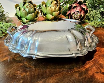 May include: A silver-plated serving tray with a scalloped edge and two handles. The tray reflects the surrounding objects, including green artichokes and foliage. The tray is on a dark wooden surface.