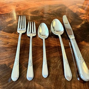 Könnte beinhalten: Ein Satz silberfarbenes Besteck, bestehend aus zwei Gabeln, zwei Löffeln und einem Messer, angeordnet auf einer Holzoberfläche. Die Utensilien haben dekorative Blumenmuster auf den Griffen. Die Löffel sind oval geformt, und das Messer hat eine gezahnte Kante.