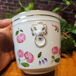 May include: White ceramic container with gold trim and a lion's head handle. The container is decorated with pink cherries and green leaves. A decorative piece for the home.