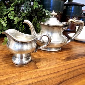 May include: A silver tea set, comprising a teapot and creamer, is displayed on a wooden surface. The teapot features a lid and curved handle, and the creamer has a spout and handle. Green foliage is in the background.