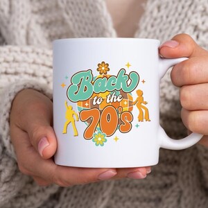 May include: A white ceramic mug featuring a colorful graphic with the text Back to the 70's. The design includes a central disco ball, two dancing figures in yellow, and small flower and star accents in shades of orange, teal, and yellow.
