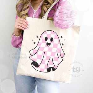 A closeup of a woman holding a tote bag leaning against a wall and the pink checkered ghost is on the front of the tote bag.