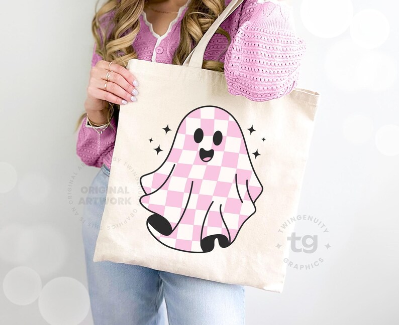 A closeup of a woman holding a tote bag leaning against a wall and the pink checkered ghost is on the front of the tote bag.