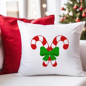 Puede incluir: Almohada blanca con un dise&ntilde;o de bast&oacute;n de caramelo rojo y blanco y un lazo verde. La almohada est&aacute; en un sof&aacute; blanco con una almohada roja detr&aacute;s.