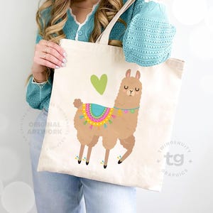 A closeup of a woman holding a tote bag leaning against the wall. The tote bag is front and center and features one of the llamas with a little green heart on the front.