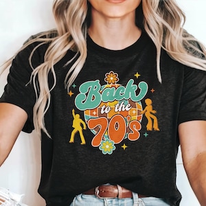 May include: A black short-sleeved t-shirt featuring a graphic print with the text Back to the 70's in teal and orange. The design includes a central disco ball, two yellow dancing figures, and small flower and star accents in various colors.