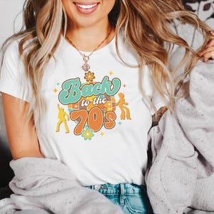 May include: A white short-sleeved t-shirt featuring a graphic print with the text Back to the 70s in teal and orange. The design includes a disco ball, two dancing figures in silhouette, and small flower icons in yellow, teal, and orange colors.