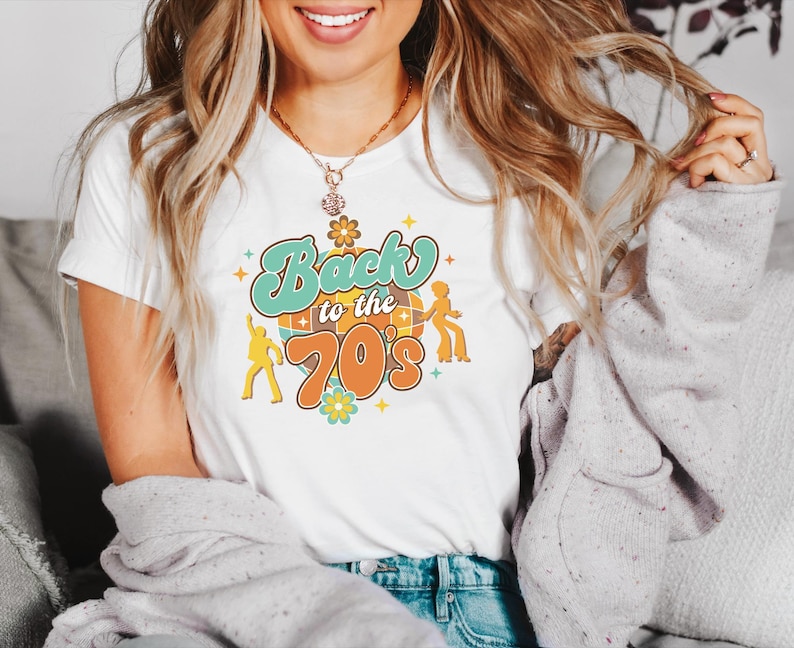 May include: A white short-sleeved t-shirt featuring a graphic print with the text Back to the 70s in teal and orange. The design includes a disco ball, two dancing figures in silhouette, and small flower icons in yellow, teal, and orange colors.
