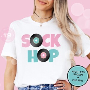 Puede incluir: Una camiseta blanca de manga corta con el texto SOCK HOP en letras de bloque rosas y turquesas. La letra O de SOCK est&aacute; sustituida por un disco de vinilo negro con el centro turquesa, y la O de HOP por un disco negro con el centro rosa.