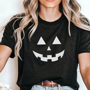 K&ouml;nnte beinhalten: Dunkelgraues T-Shirt mit einem wei&szlig;en K&uuml;rbisgesicht. Das Design umfasst dreieckige Augen, eine dreieckige Nase und ein breites, zahnreiches Grinsen. Das Shirt ist ein Rundhals-Stil.