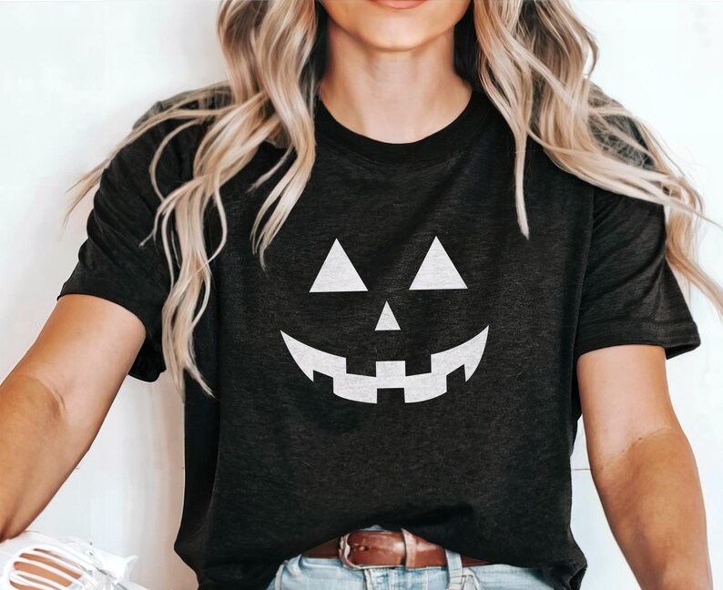 K&ouml;nnte beinhalten: Dunkelgraues T-Shirt mit einem wei&szlig;en K&uuml;rbisgesicht. Das Design umfasst dreieckige Augen, eine dreieckige Nase und ein breites, zahnreiches Grinsen. Das Shirt ist ein Rundhals-Stil.